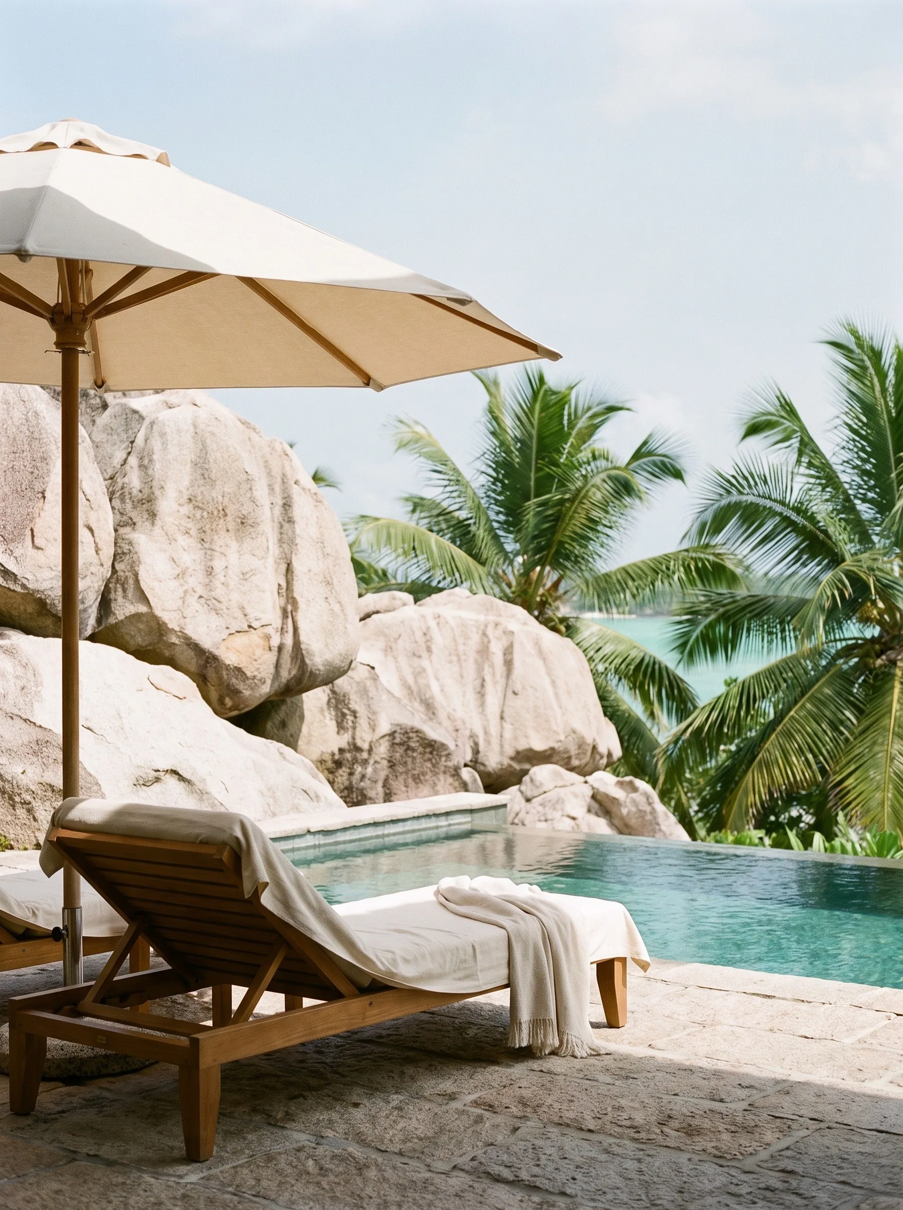 A sun lounger and parasol beside a pool among granite boulders and palms